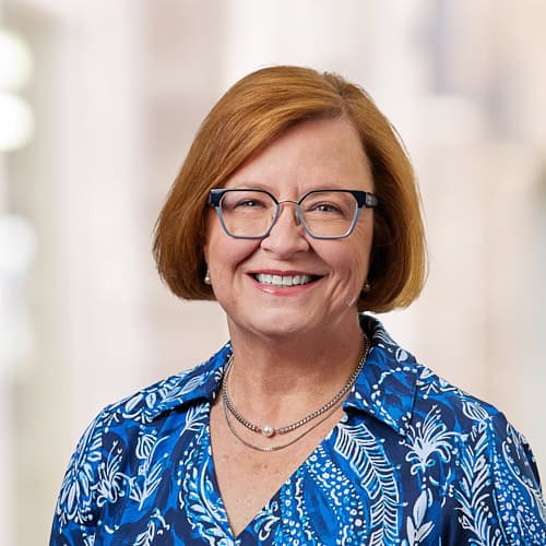 Cindy L. Corpier, MD, a woman with shoulder-length red hair and glasses, smiles at the camera. She wears a blue patterned blouse and a pearl necklace. The background is softly blurred, focusing on her cheerful expression.