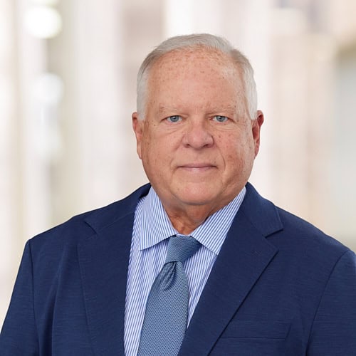 An older man, possibly Mr. Kyle Smith, with short white hair is wearing a navy blue suit, light blue and white striped shirt, and a light blue tie. He poses against a blurred indoor backdrop.