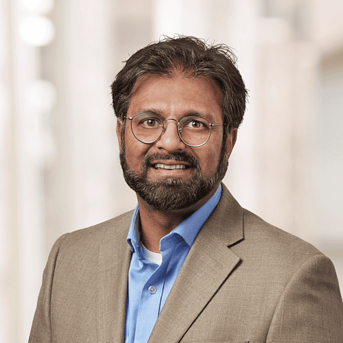 A man with glasses and a beard, dressed in a beige blazer and blue shirt, smiles while posing in a brightly lit indoor setting with blurred background elements. This image of Muhammad Omar Azam, MD captures his warm demeanor perfectly.