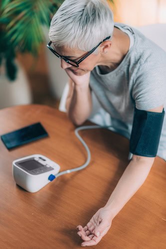 A person with short gray hair and glasses is seated at a table, using a digital blood pressure monitor. Their left arm, likely recovering from a recent kidney transplant, has a cuff attached while they lean their head on their right hand. A smartphone is placed on the table beside them.