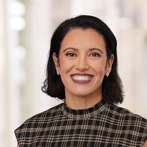 Cristina Arce, with an MD and MSc, is pictured smiling against a blurred background, her dark hair complementing a checkered top.