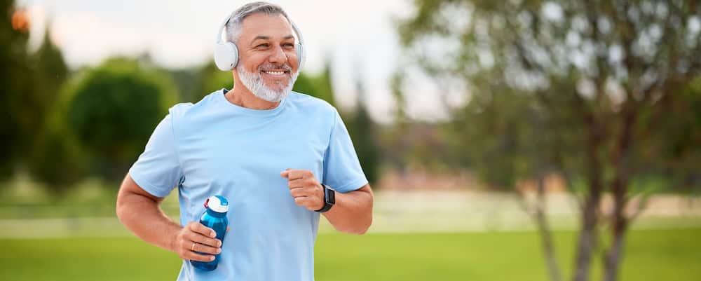 A smiling man with gray hair and a beard, who recently underwent a successful kidney transplant, jogs in the park while wearing headphones and a light blue shirt. He holds a water bottle in one hand. The background is green and blurred, suggesting trees and grass.