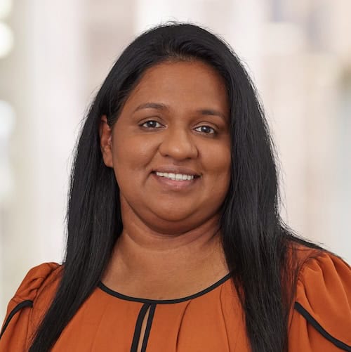 Jeena Koshy, APRN, FNP-BC, with long, dark hair, smiles at the camera. She wears an orange top with black trim against a softly blurred background.