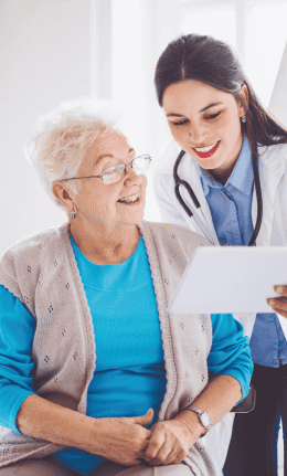 A healthcare professional wearing a stethoscope shows a document on diet and nutrition to an elderly woman. The woman, smiling and wearing glasses and a blue shirt with a beige cardigan, is in a brightly lit room, attentively listening. The purpose of this image is to demonstrate how easy it is to discuss Chronic Kidney Disease with your medical provider