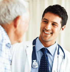 A smiling doctor with a stethoscope discusses diet and nutrition with an older man. The doctor, in a white coat, blue shirt, and dark tie, engages warmly in a medical office or clinic setting, where curtains add a touch of privacy to their conversation. The purpose of this image is to demonstrate how simple it is to discuss kidney disease with your patient. 