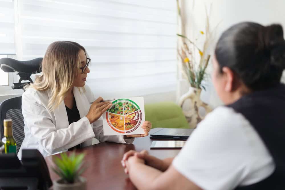 A nutritionist in a white coat explains a healthy eating chart to a seated person living with chronic illness. The chart shows different food groups crucial for managing chronic kidney disease. They're in an office setting with plants and large windows, sharing stories of hope and healing strategies.