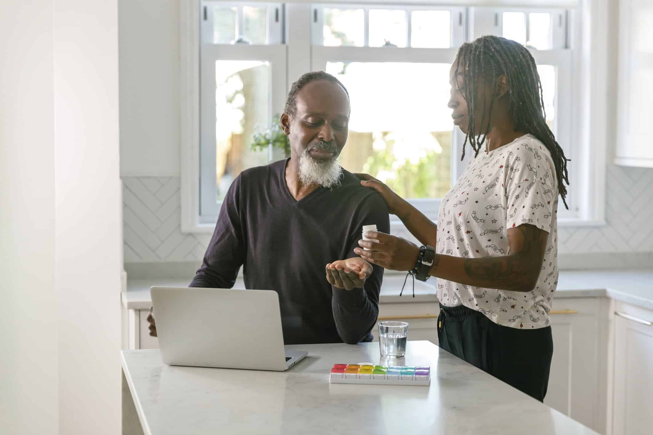 A man recovering from a kidney transplant sits at a kitchen counter with his laptop, taking a pill handed to him by a woman beside him. A glass of water and a pill organizer are on the counter, illuminated by the room's bright natural light.