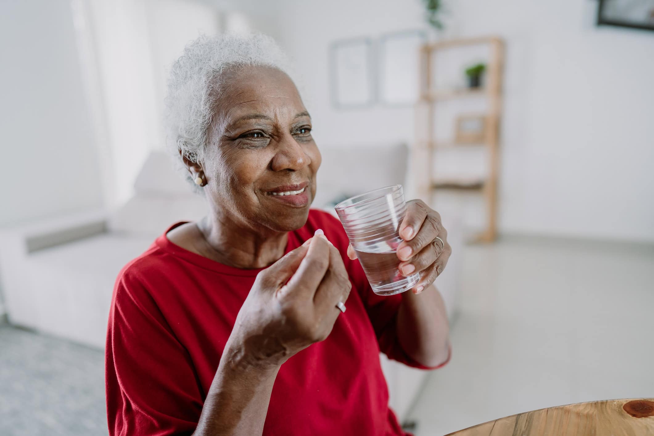 An elderly woman with short gray hair sits in a bright room, smiling in her red shirt as she holds a glass of water and a pill, part of her weight management routine. In the background, a blurry bookshelf adds to the cozy atmosphere.