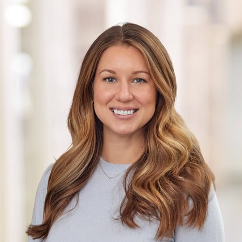 Erin Himes, AGNP-C, is smiling with long, wavy brown hair cascading over her shoulders. She wears a light gray top and a delicate necklace. The softly blurred background hints at an indoor setting, adding to the serene ambiance of this APRN and MSN professional's portrait.