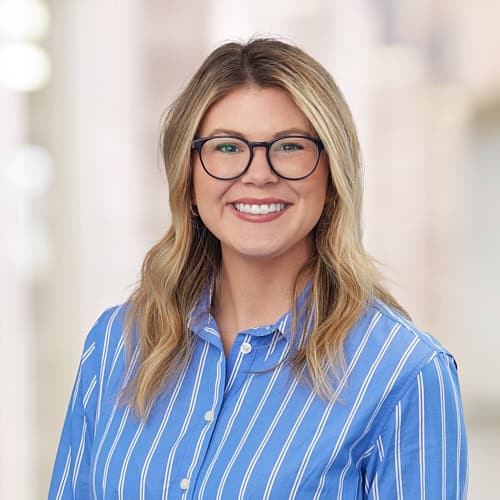 Erin Himes, MSN, AGNP-C, beams beneath her long blonde hair and black-framed glasses. Her blue shirt with white vertical stripes complements the softly blurred indoor setting, offering a glimpse into the professional warmth of this APRN.