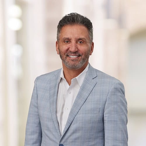 Hassan N. Ibrahim, a distinguished MD with gray hair and beard, smiles warmly at the camera in his light blue checked blazer and white shirt. The blurred background draws attention to his friendly demeanor.